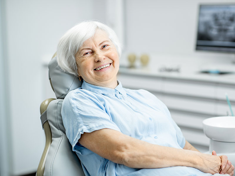 An elderly woman is seated in a dental chair, smiling at the camera.