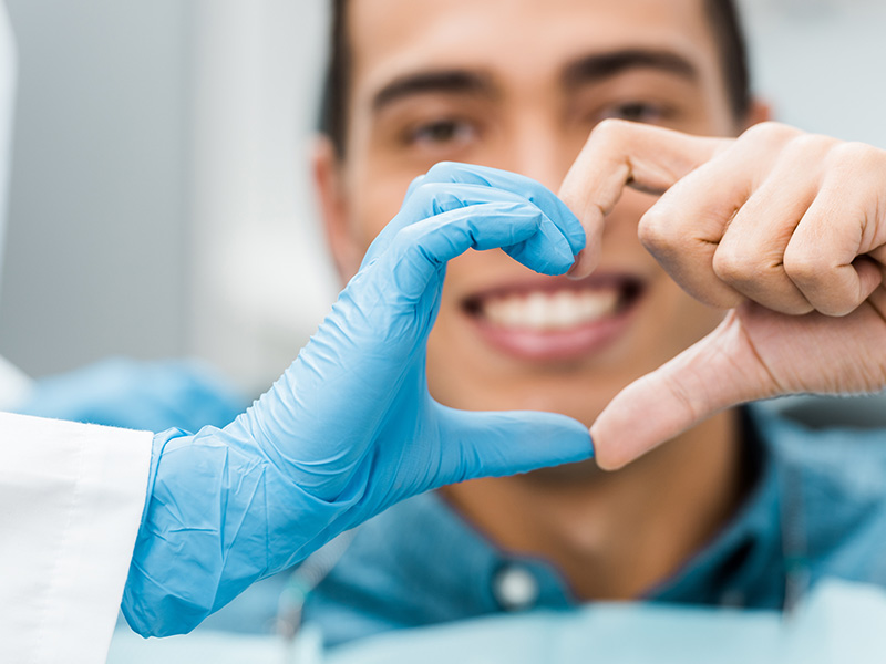 A smiling person wearing a blue surgical mask and gloves is holding up two fingers in a heart shape, with another individual in the background also wearing a blue surgical mask and holding their hand near the first person s hand.